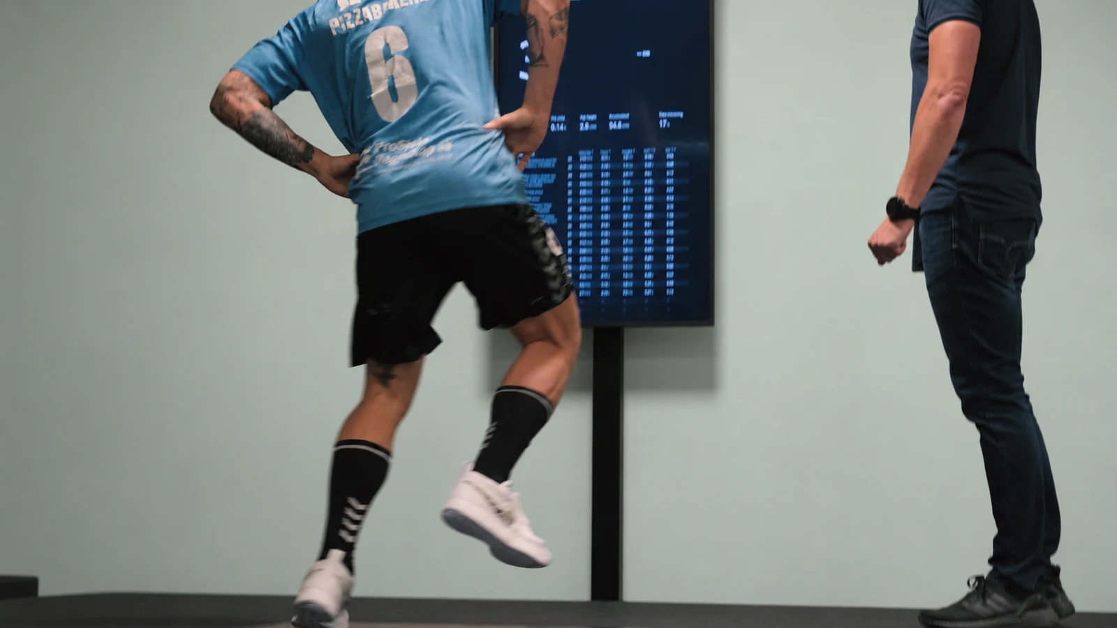 A person in a blue jersey performs an exercise in front of the Alphatek display while a professional observes.