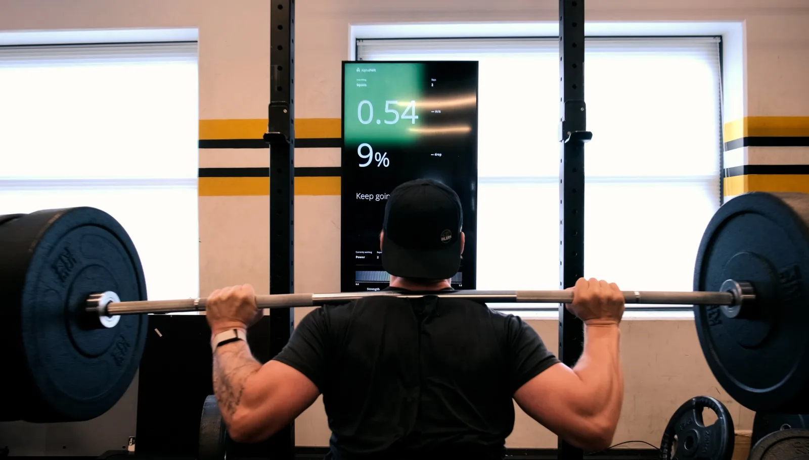An athlete with a barbell on the shoulders in front of the Alphatek display showing 0.54 m/s and a 9 percent velocity drop.