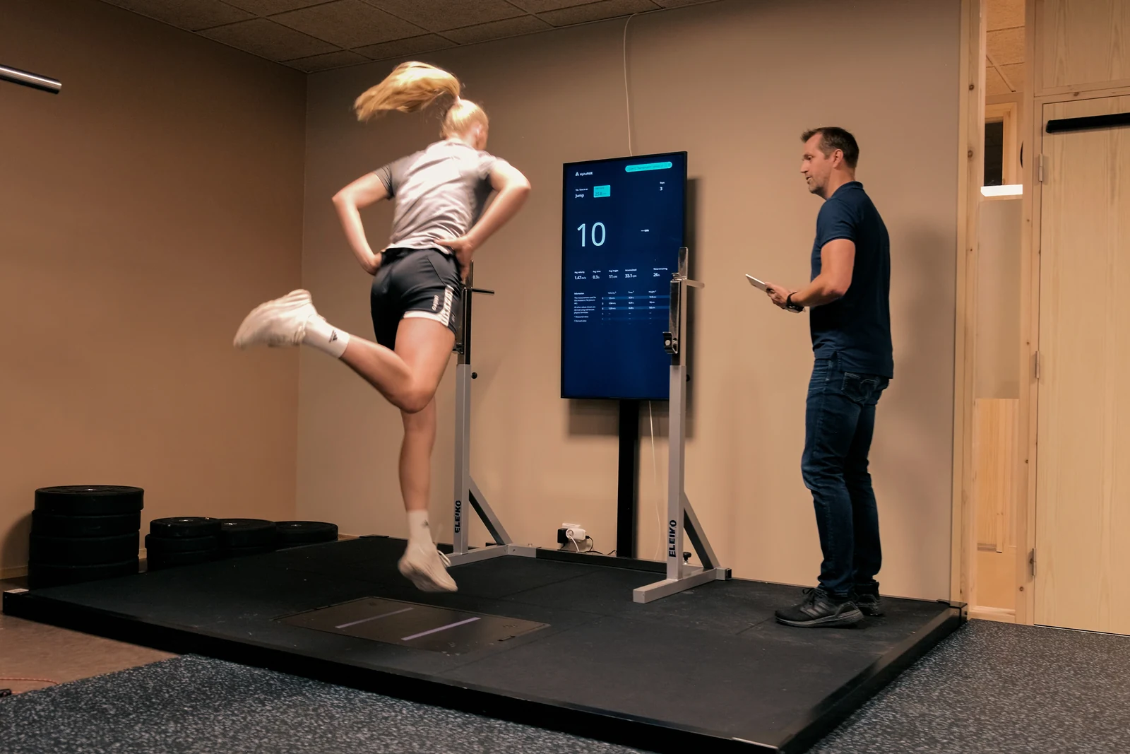 A participant hops on one leg on the Alphatek platform while a guide observes with a tablet.