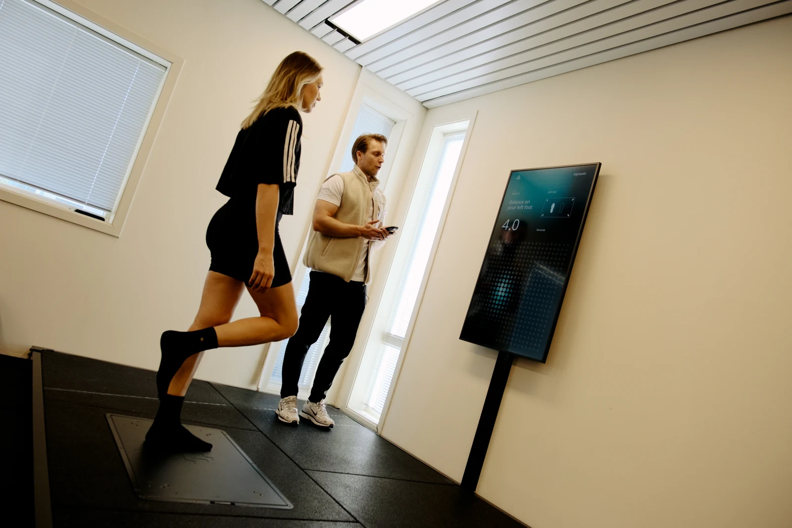 A member balances on one leg on the Alphatek platform while a trainer guides the activity beside the display.
