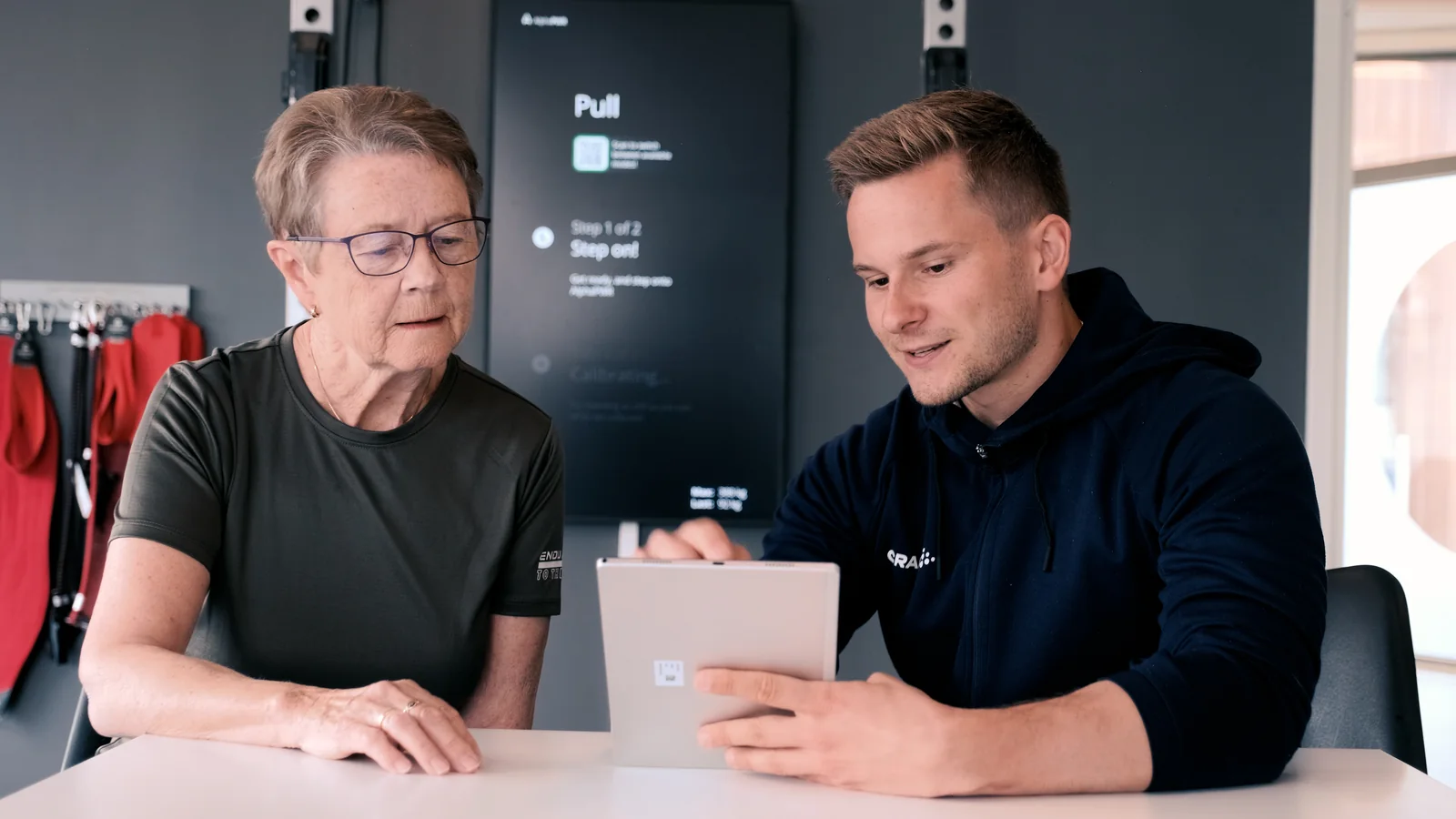 Two people review a tablet together, with the Alphatek display in the background.