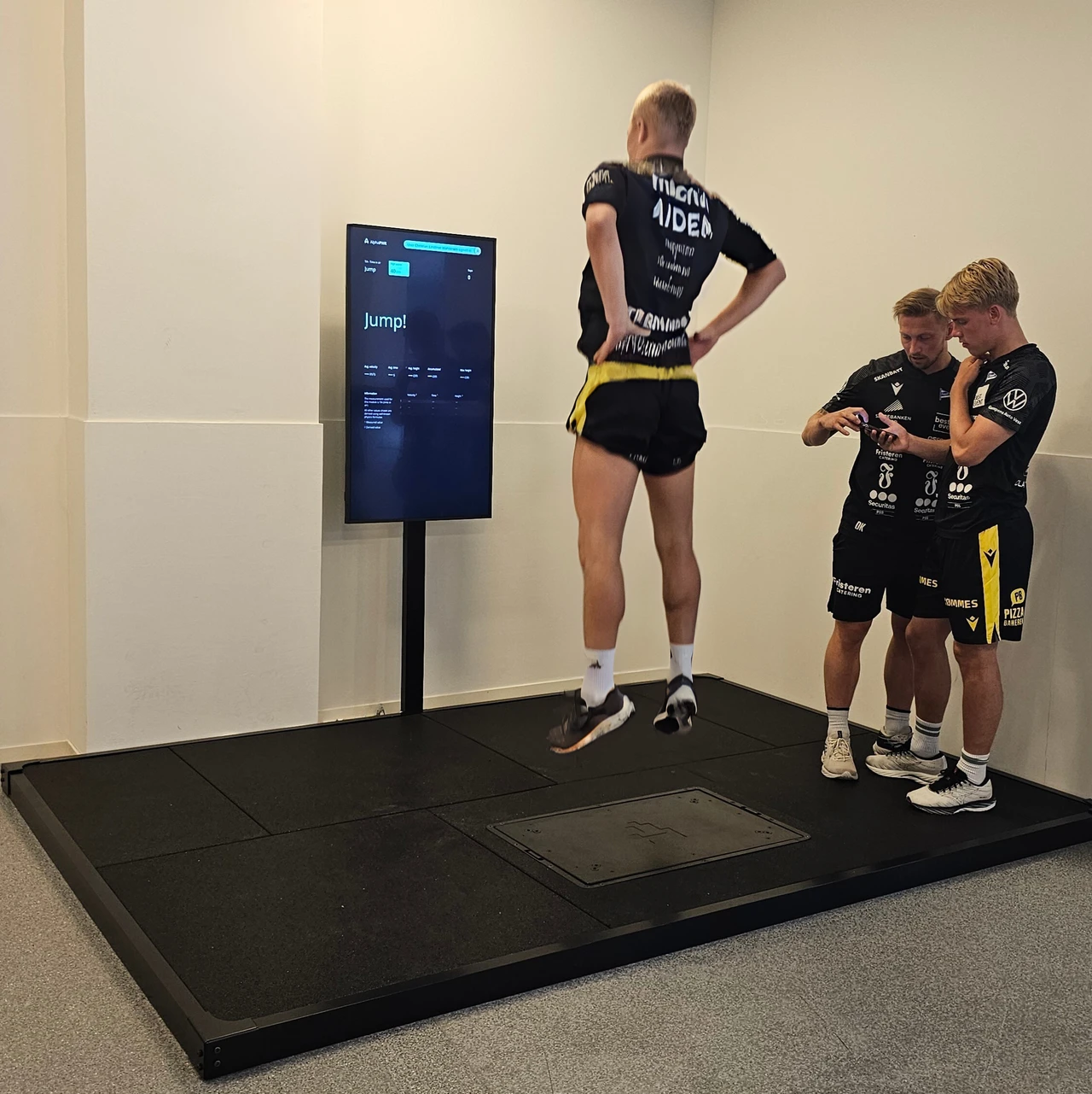 An athlete jumps on the Alphatek platform while two others review a phone beside him.