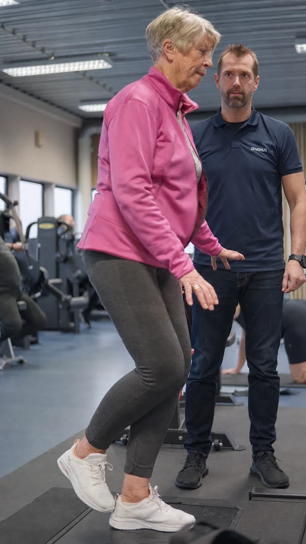 An older woman balances on one leg on the Alphatek platform, with a trainer beside her.