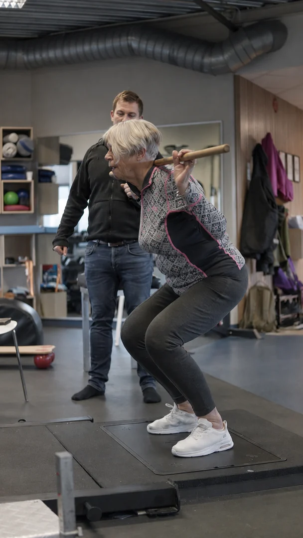An older woman performs a squat with a bar across her shoulders on the Alphatek platform, with a trainer behind her.